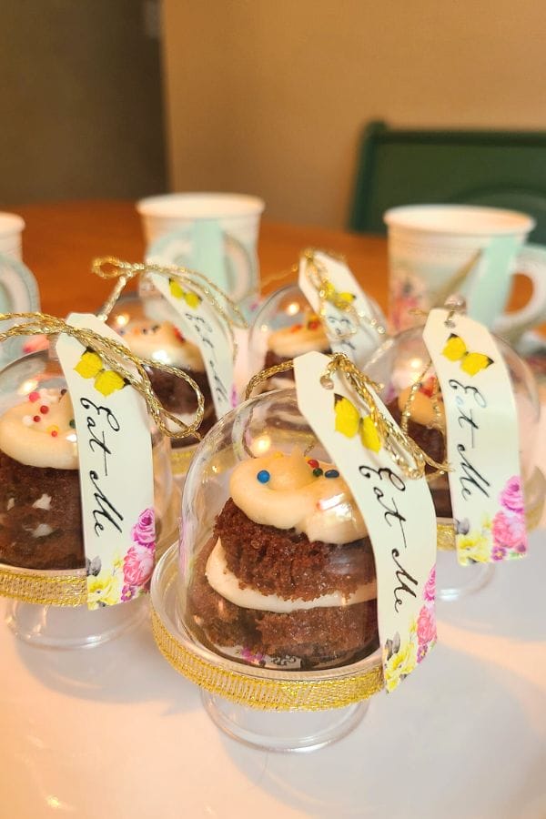 Mini cakes in clear domes with gold ribbons and tags, decorated with frosting and sprinkles, add a sweet touch to a Classic Literature Themed Tea Party table adorned with patterned cups.