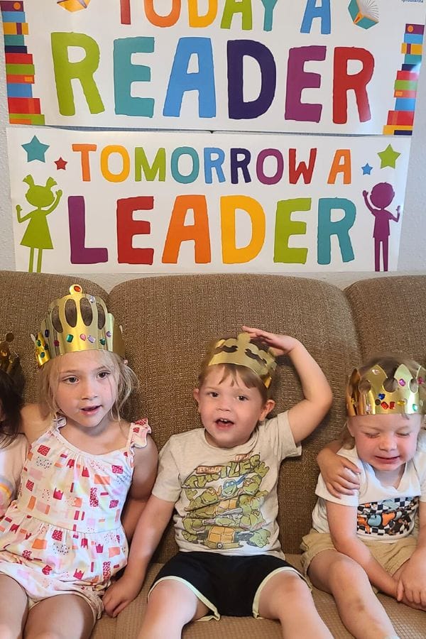 Three children wearing paper crowns sit on a couch in front of a colorful sign that reads "Today a Reader, Tomorrow a Leader," celebrating their Classic Literature Themed Tea Party.