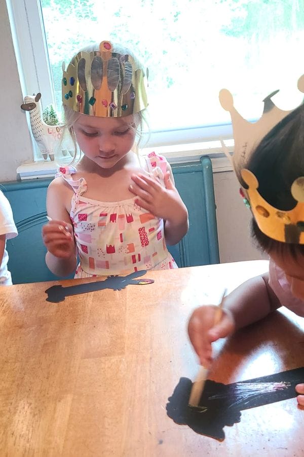 In a bright room, two children wearing paper crowns paint black shapes on a wooden table, setting the stage for a classic literature-themed tea party.