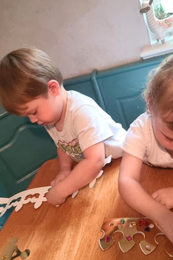Two children are sitting at a table, piecing together a cardboard crown shape decorated with colorful stickers, envisioning themselves as characters in a classic literature-themed tea party.