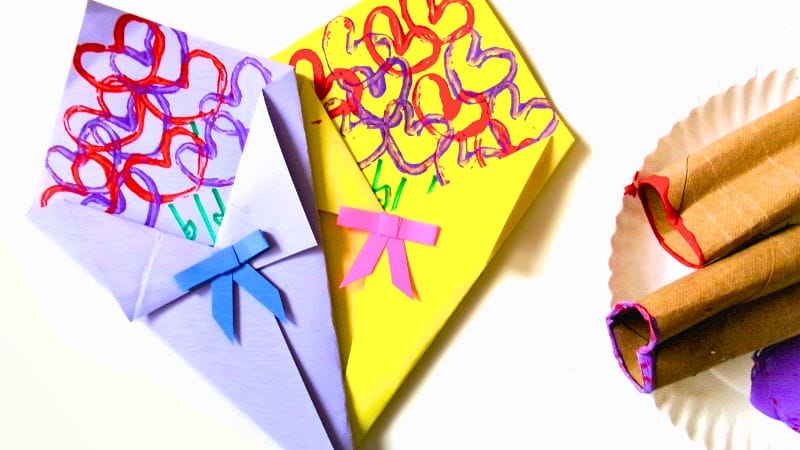 Two colorful handmade envelopes with heart patterns and bows on a white surface; a charming bouquet of heart stamps accompanies paper tubes and a plate nearby.