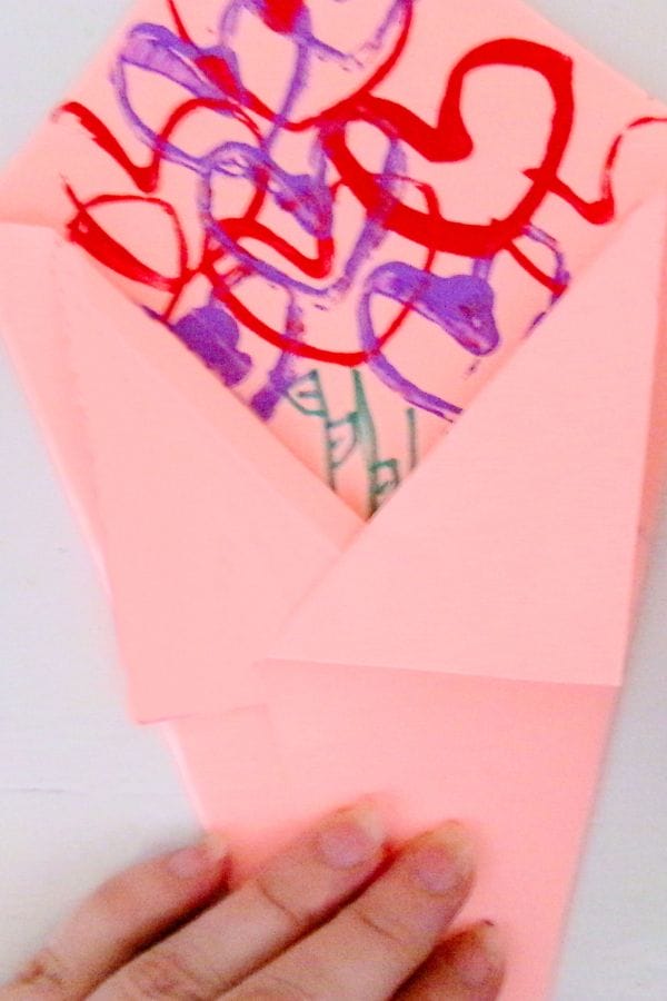 A hand holds a pink, heart-patterned paper wrapped like an envelope, adorned with a delicate bouquet illustration that seems almost ready for a heart stamp to seal it with love.