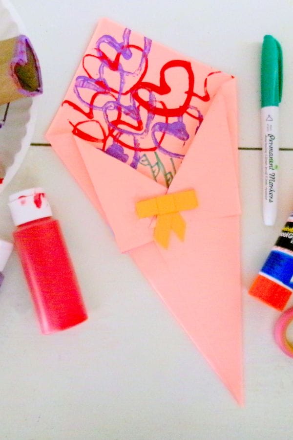 A pink origami envelope adorned with colorful scribbles sits surrounded by red paint, a green marker, and glue sticks on the table. Nearby, a bouquet of heart stamps adds an extra touch of charm to this creative scene.