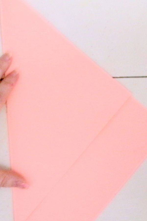 A hand expertly folds pink paper into a triangular shape, as if crafting the foundation for a heart stamp design, on a pristine white surface.