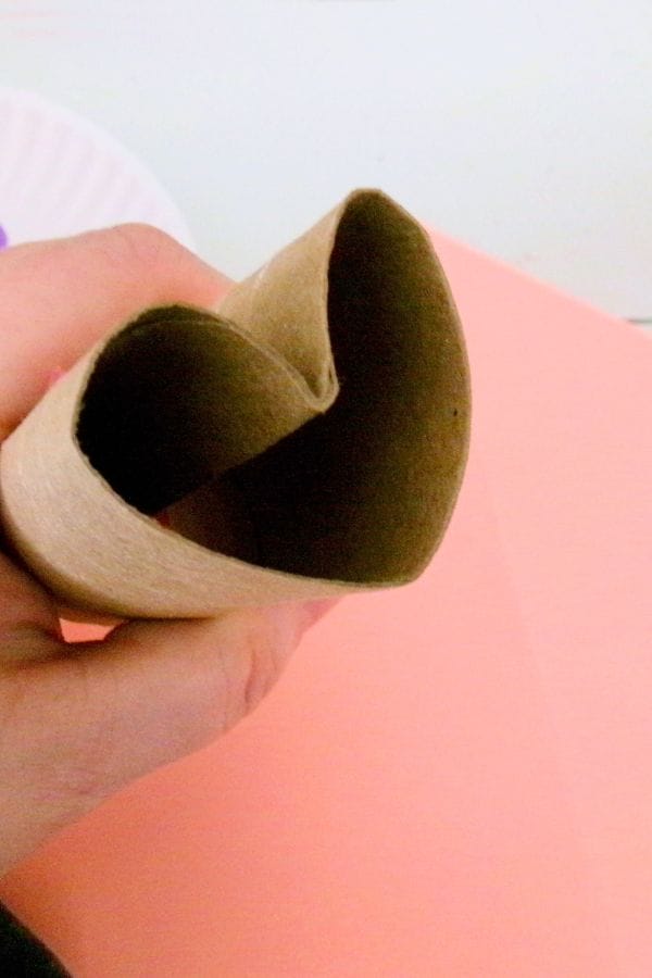 A hand holds a cardboard tube shaped into a heart, like a stamp ready to imprint love on paper, against a soft pink background.