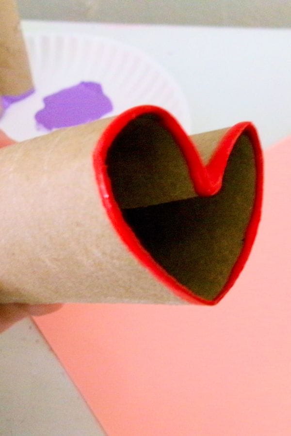 Cardboard tube shaped into a heart with red-painted edges, resembling a heart stamp, held against a bouquet-like backdrop of pink and purple papers.
