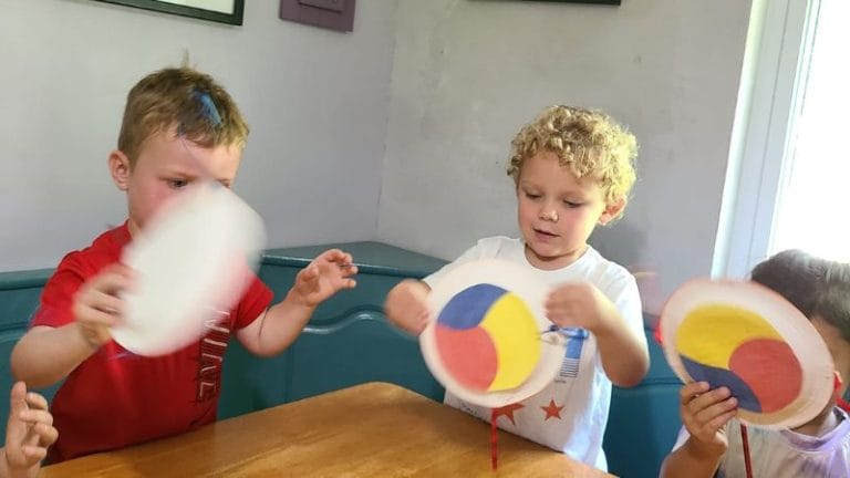 Children sitting at a table, holding paper plates with multicolored circles, engaging in a craft activity.