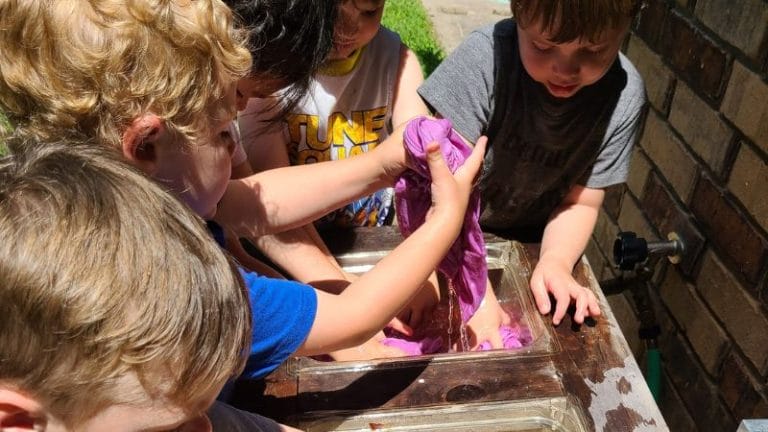 Children engaged in creative play with purple slime outdoors at a water table near a brick wall, subtly inspired by lesson plans on Native American culture, fostering both fun and learning in an imaginative setting.