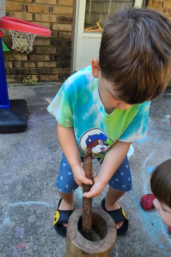 A child in a tie-dye shirt uses a stick to grind something in a wooden mortar outdoors, reminiscent of lesson plans on Native American traditions. In the background, a basketball hoop stands beside colorful chalk drawings, adding charm to this playful scene.