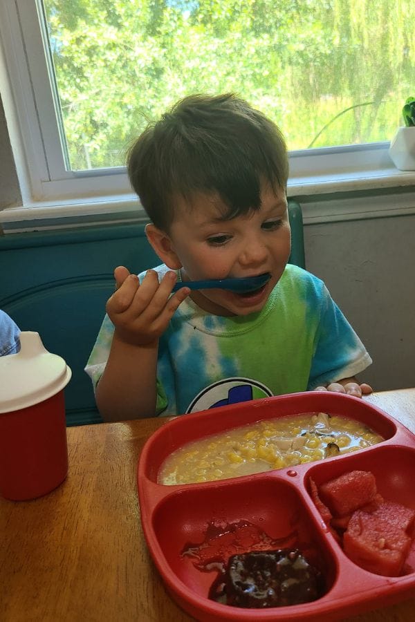 A child in a tie-dye shirt eats with a blue spoon from a divided red plate, reminiscent of lesson plans on Native American culture's vibrant colors. A red cup sits nearby, while a window frames the lush greenery outside.