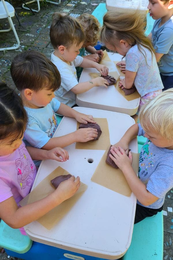 Children sitting at tables outdoors, engaged in crafting with clay as part of lesson plans on Native American art and culture.