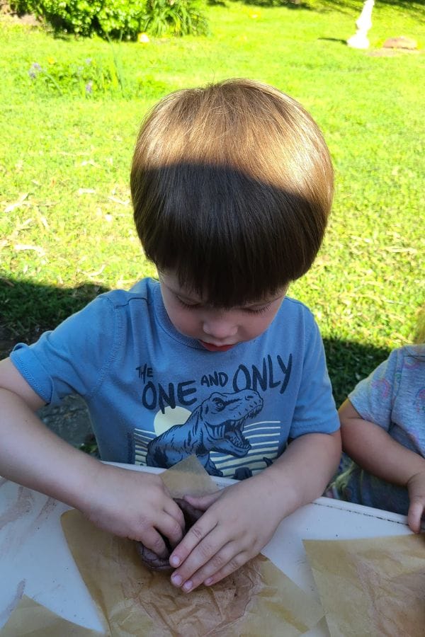 A child in a blue dinosaur shirt explores the outdoors with clay on a sunny day, inspired by lesson plans on Native American art and culture.