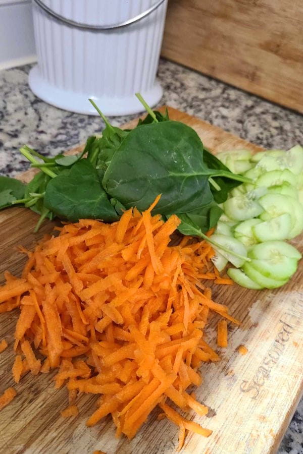 Shredded carrots, spinach leaves, and sliced cucumbers lay invitingly on a wooden cutting board in a kitchen setting, ready to be transformed into a vibrant tuna wrap without tortillas.