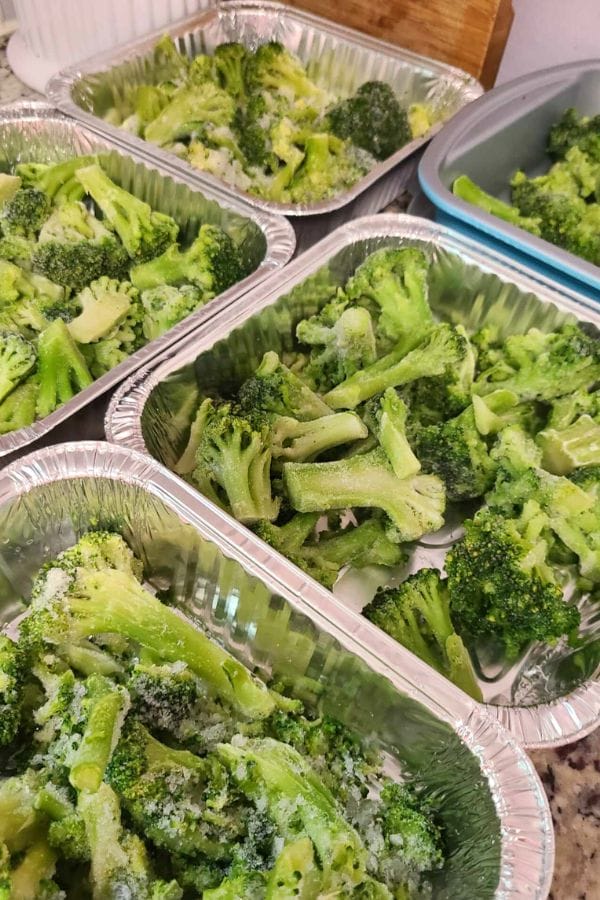 Several aluminum trays filled with frozen broccoli florets and the ingredients for a jalapeno popper chicken casserole are displayed on a kitchen counter.