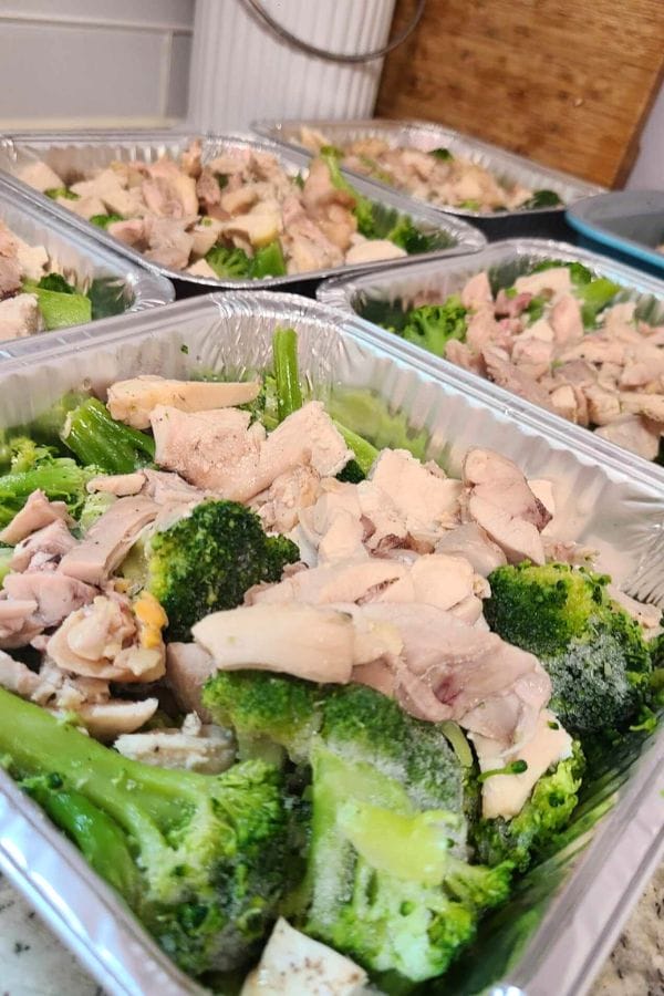 Aluminum trays filled with Jalapeno Popper Chicken Casserole, featuring cooked broccoli and chicken pieces, arranged in rows on a kitchen counter.