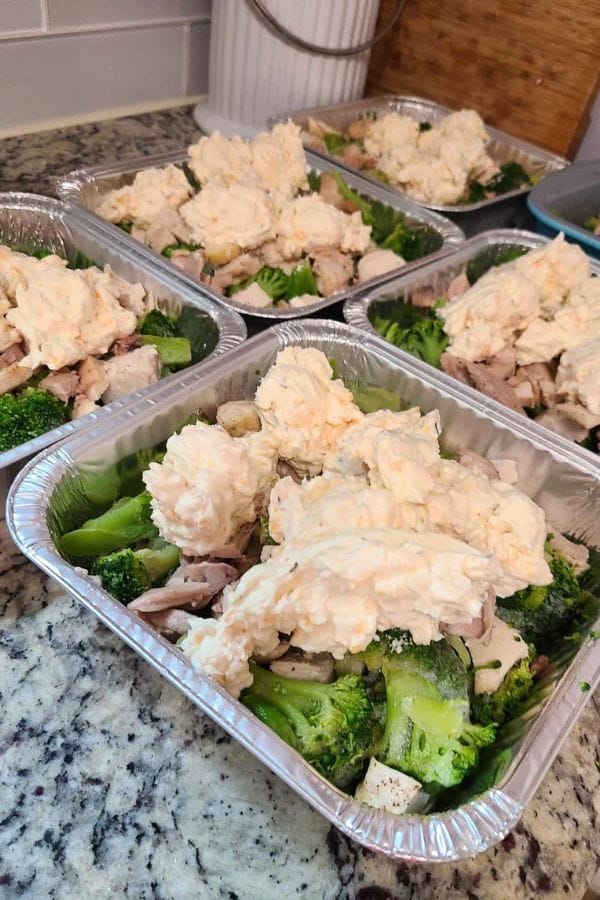 Five foil containers filled with broccoli, chicken, and a dollop of cheese mix, resembling a delicious Jalapeno Popper Chicken Casserole, are neatly placed on a granite countertop.