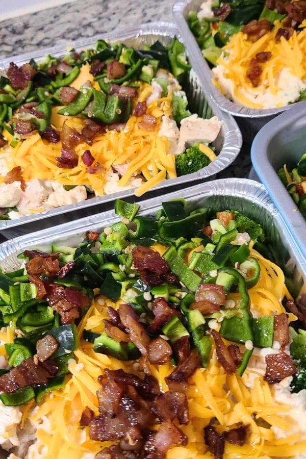 Close-up of three foil trays filled with Jalapeno Popper Chicken Casserole, topped with chopped bacon, shredded cheese, green peppers, and onions.