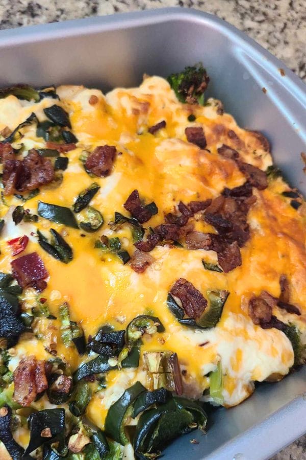 Savor the zesty Jalapeno Popper Chicken Casserole, a baked dish with melted cheese, bacon, green vegetables, and fiery peppers in a metal pan.