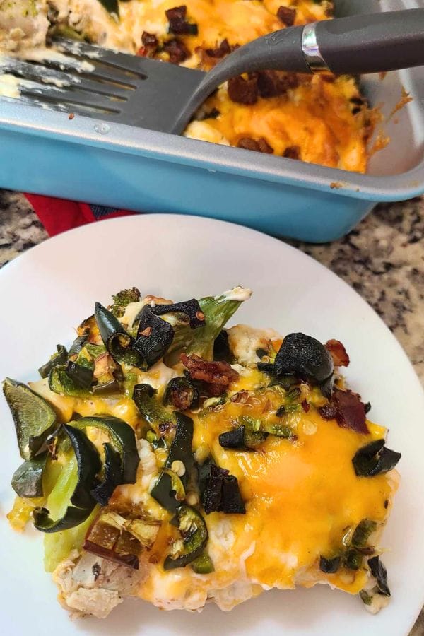 A plate of cheesy jalapeno popper chicken casserole with vegetables, including broccoli and olives, topped with melted cheese, next to a blue baking dish with a serving spatula.