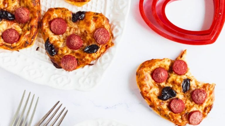 Mini heart-shaped pizzas topped with pepperoni and olives sit elegantly on white plates, accompanied by a red heart-shaped mold and gleaming forks.