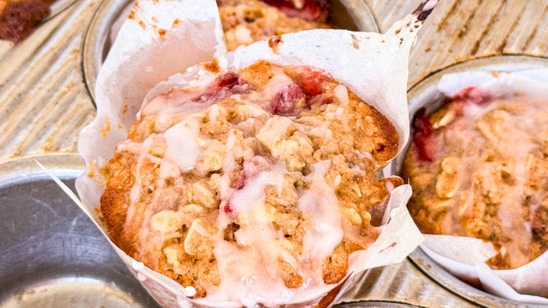Close-up of strawberry oatmeal muffins with pink icing and crumb topping, wrapped in parchment paper, nestled perfectly in a muffin tin.