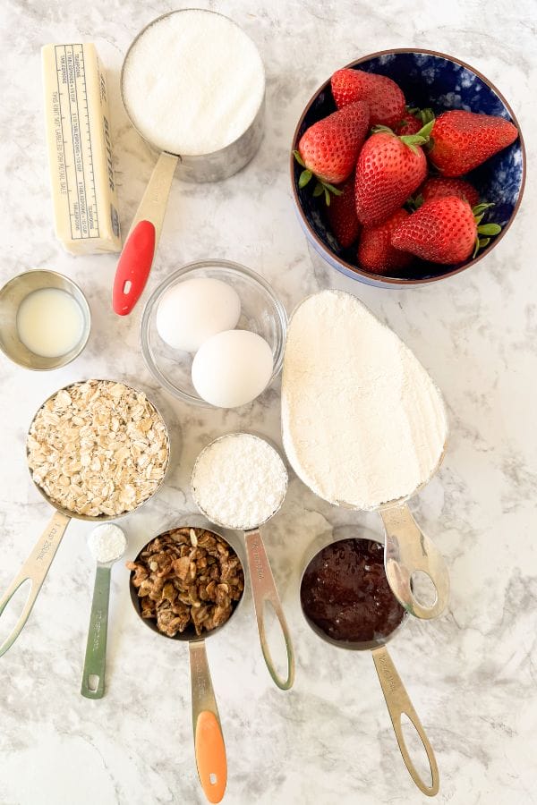 Top-down view of ingredients for strawberry oatmeal muffins: strawberries, sugar, butter, eggs, flour, oats, walnuts, vanilla, baking soda, and jam are neatly placed in separate measuring cups and bowls on a marble surface.