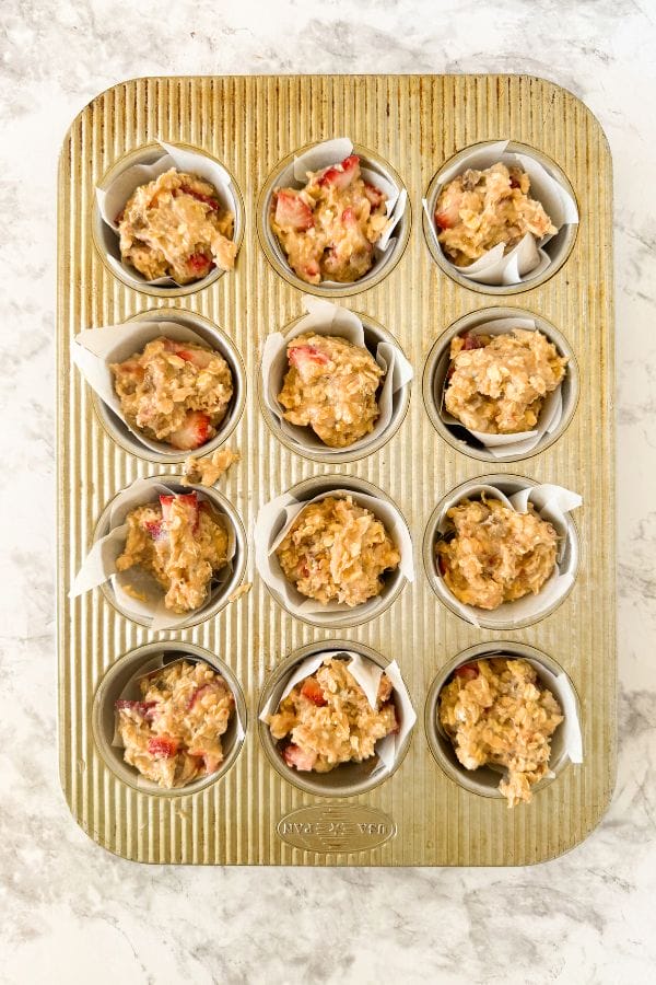 A muffin tin with twelve slots, each filled with strawberry oatmeal muffin batter, ready to be baked. The batter contains visible chunks of apple and oats. The tin sits on a marble countertop.