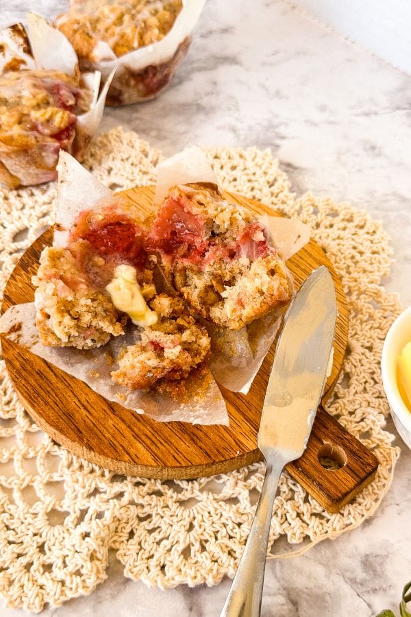 A warm strawberry oatmeal muffin with a pat of melting butter is cut open on a wooden board, accompanied by a silver butter knife. In the background, more muffins rest gracefully on a lace doily.