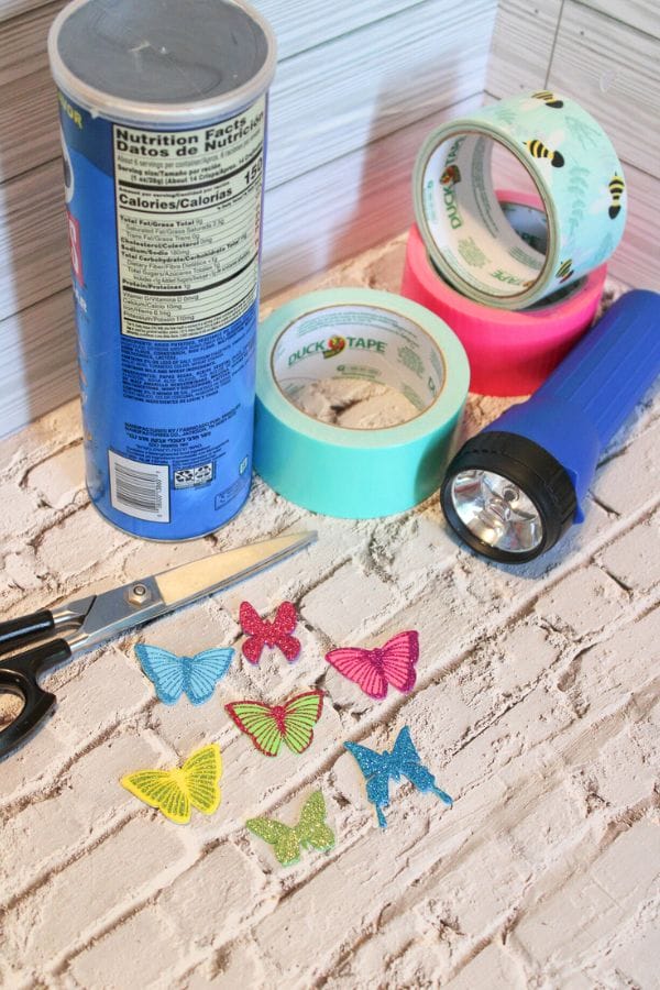 Craft supplies for DIY shadow puppets include a can, assorted decorative tapes, a blue flashlight, scissors, and colorful butterfly cutouts on a textured surface.