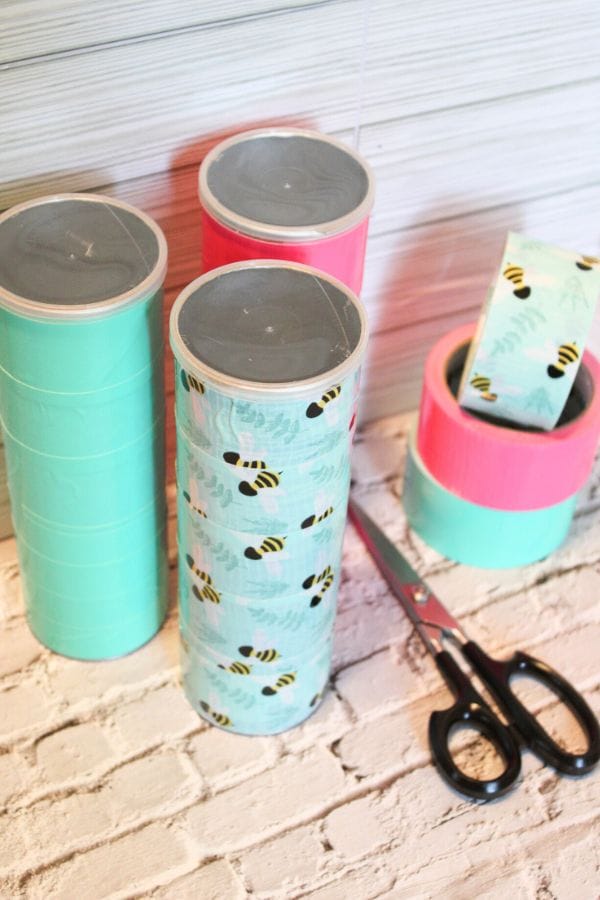 A vibrant collection of colorful duct tape rolls, featuring charming bee patterns, is stacked on a white brick surface next to a pair of scissors. Perfect for crafting projects like DIY shadow puppets, this setup inspires creativity and imagination in any craft enthusiast.
