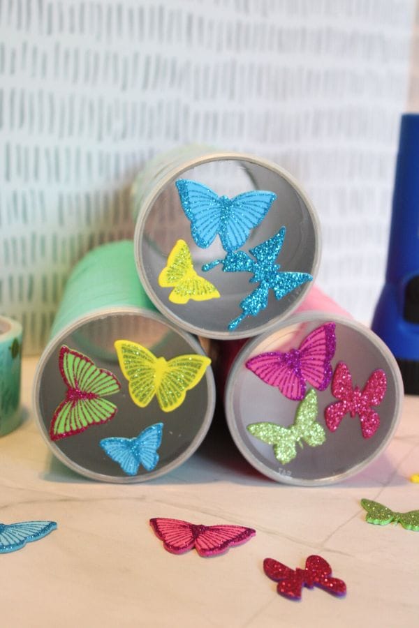 Three rolls of tape stand on their ends, each adorned with colorful glittery butterfly stickers, ready to become the base for a spontaneous DIY shadow puppets project.