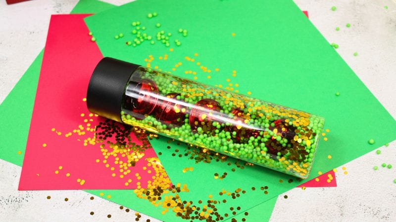 A Winter Sensory Bottle adorned with green and yellow beads and red ornaments lies elegantly on festive red and green paper.