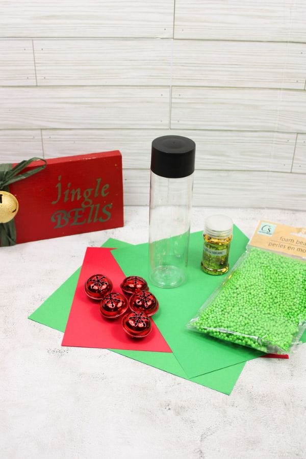 Craft supplies for Winter Sensory Bottles include a clear bottle with a black cap, red bells, a small jar of glitter, green beads, red and green paper, and a red "Jingle Bells" sign on a white surface.