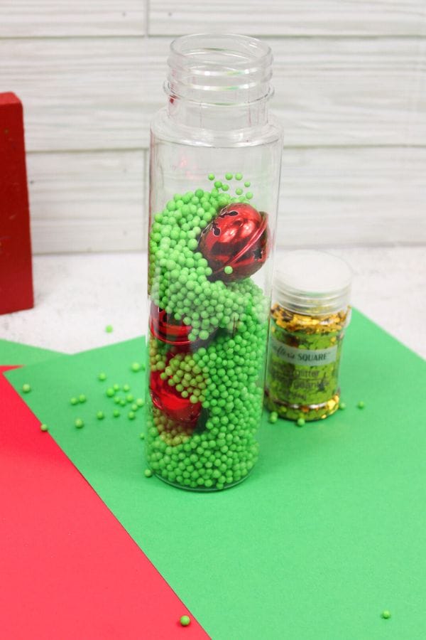 A clear bottle filled with green beads and red jingle bells evokes the festive charm of Winter Sensory Bottles. Nearby, a small jar of gold sequins completes the scene on a red and green background.