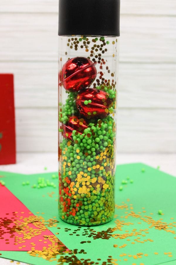 A clear container, perfect as a Winter Sensory Bottle, is filled with green beads, red metallic bells, and gold confetti, set against a backdrop of festive red and green paper.