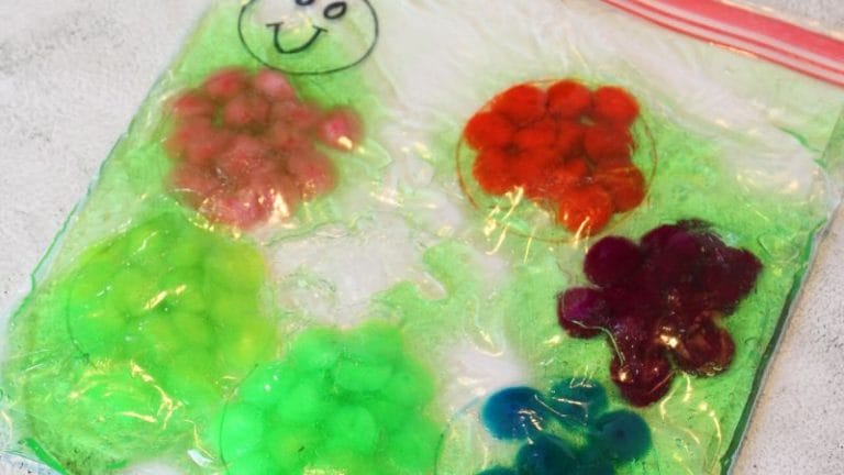 A clear ziplock bag, resembling a caterpillar sensory bag, is filled with colorful blobs of slime in red, green, purple, and blue on a light surface. A smiley face is drawn on the bag.