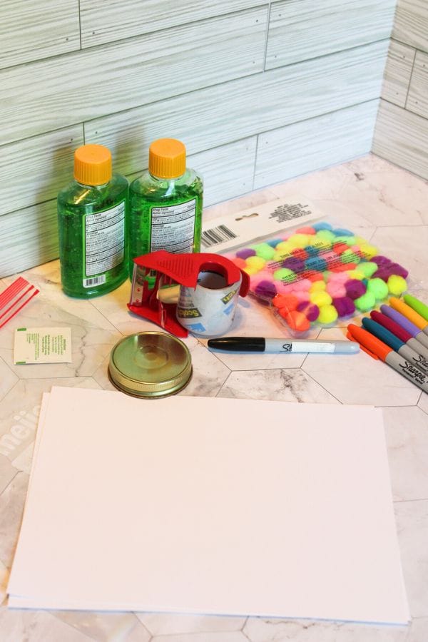 A set of craft supplies on a table, perfect for creating a Caterpillar Sensory Bag, includes bottles of green liquid, a pack of pom-poms, tape, paper, markers, and a jar lid.