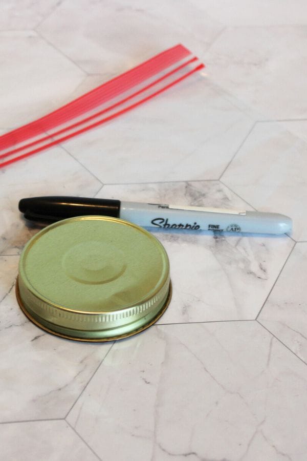 A marker, a metal jar lid, and several red plastic strips are arranged on a white, marble-patterned surface, reminiscent of the vibrant textures in a Caterpillar Sensory Bag.