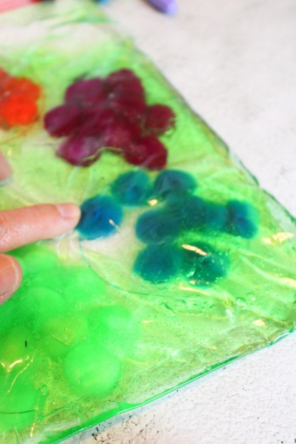 A person's finger presses on a vibrant Caterpillar Sensory Bag filled with colorful gel and objects, including blue, red, and green shapes, against a light surface.