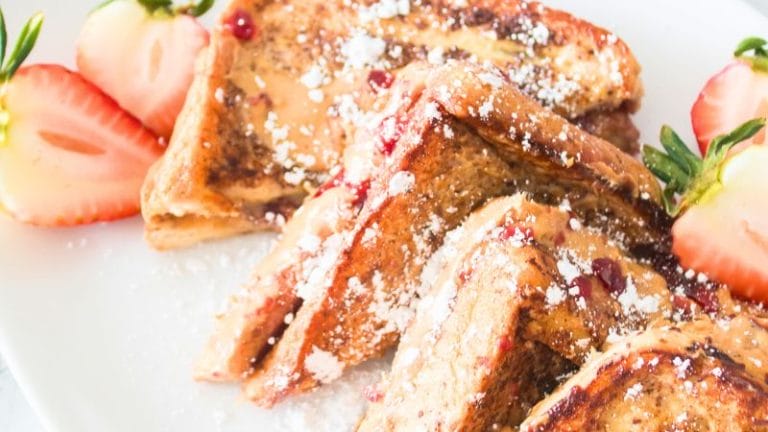 Close-up of peanut butter and jelly stuffed French toast sprinkled with powdered sugar, garnished with halved strawberries on a white plate.