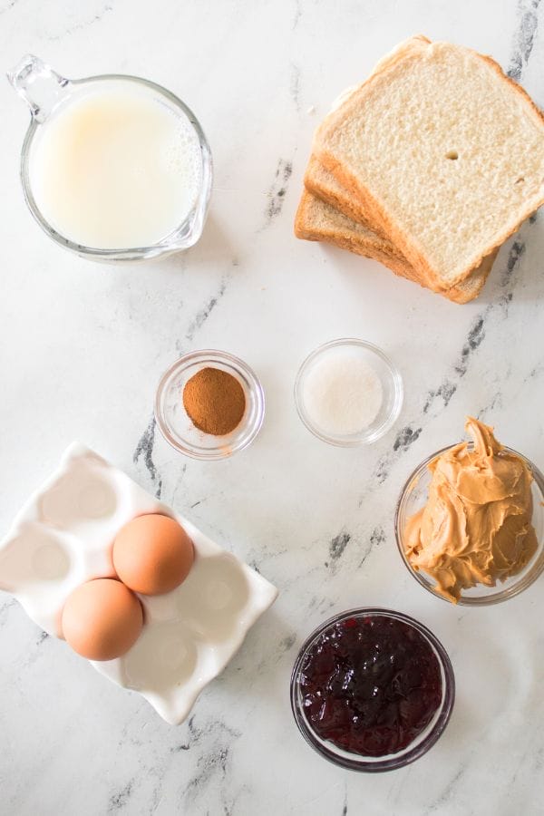 Peanut butter and jelly stuffed French toast ingredients—bread slices, milk, eggs, cinnamon, sugar—are beautifully arranged on a marble surface.