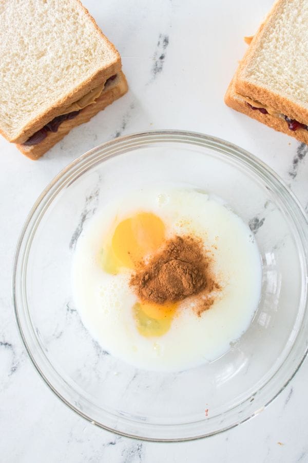 A glass bowl with milk, eggs, and cinnamon sits beside two peanut butter and jelly sandwiches on white bread, ready to be transformed into delicious Peanut Butter and Jelly Stuffed French Toast.