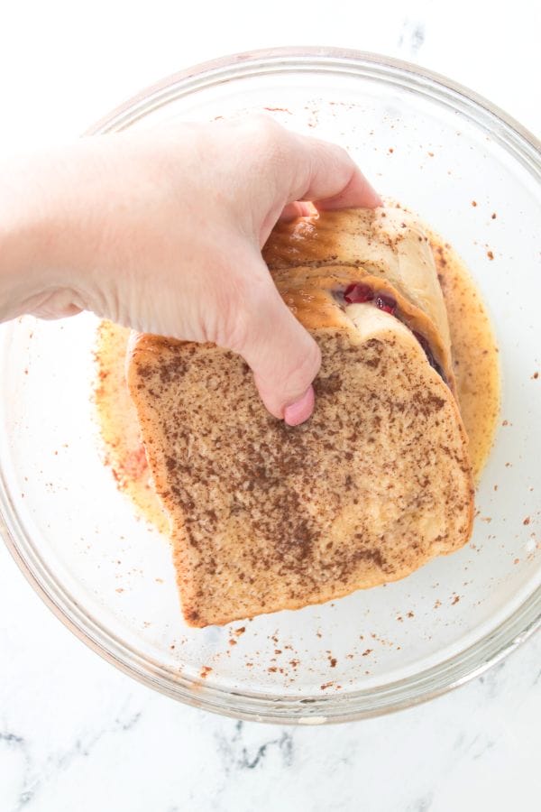 A hand dips a slice of cinnamon-swirl bread into an egg mixture in a glass bowl, preparing the base for a decadent peanut butter and jelly stuffed French toast.