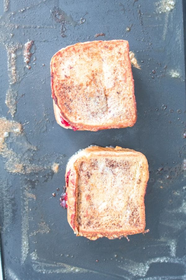 Two grilled cheese sandwiches with a hint of jam, reminiscent of a savory twist on peanut butter and jelly stuffed French toast, are on a black griddle, showing golden-brown toasted bread.