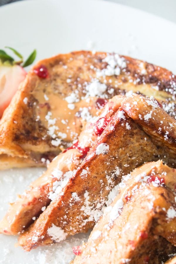 Close-up of Peanut Butter and Jelly Stuffed French Toast slices, adorned with a peanut butter and jam topping, dusted with powdered sugar, and accompanied by a strawberry garnish on the side.