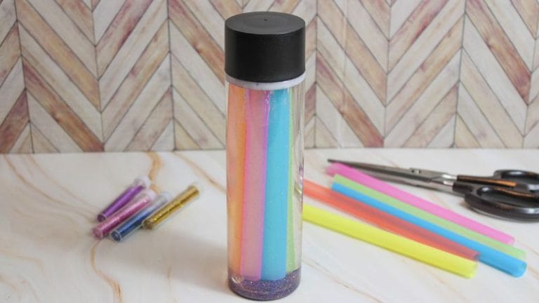 A plastic bottle, labeled "After the Storm Sensory Bottle," brims with colorful liquids and glitter on a table. Surrounding it are tubes of glitter, vibrant straws, and scissors. The backdrop showcases a chevron-patterned wall.
