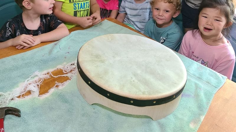 Children gathered around a table, marveling at a large round drum placed on a green cloth, as part of their lesson plans on Irish culture.