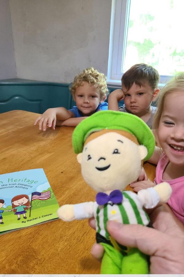 Three children sit at a table with a person holding a plush toy in the foreground. A children's book on Irish culture, part of engaging lesson plans, is spread out on the table.