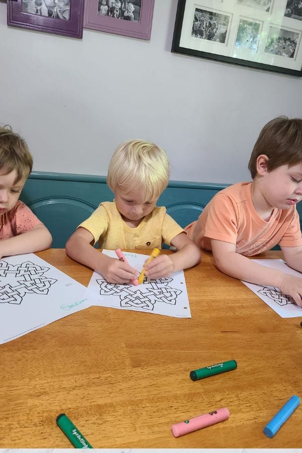 Three young children sit at a wooden table, eagerly coloring in printed knot designs with markers as part of their lesson plans on Irish culture. Framed photos adorn the wall behind them, creating a cozy backdrop for this educational activity.