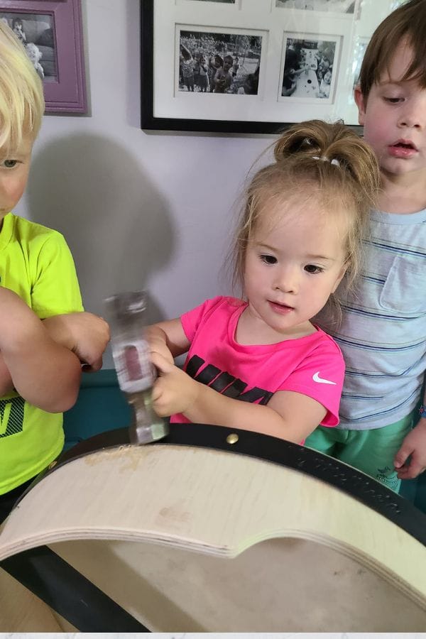 Young girl in a pink shirt uses a hammer on a wooden surface, engaged in an activity inspired by lesson plans on Irish culture, as two other children watch. Framed photos are on the wall behind them.